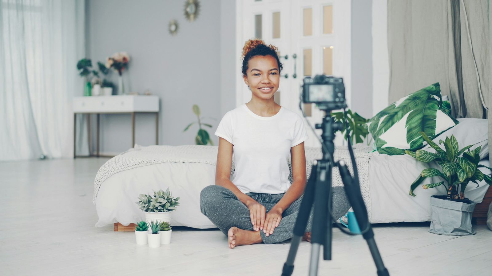 A woman sitting on the floor of a bright room, recording herself with a camera on a tripod