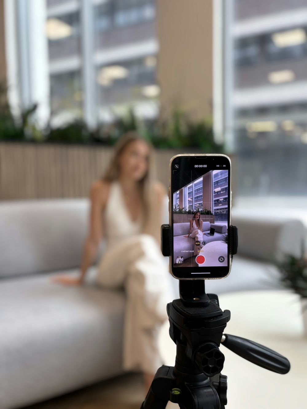 A smartphone filming a woman on a sofa from a tripod in a bright living room